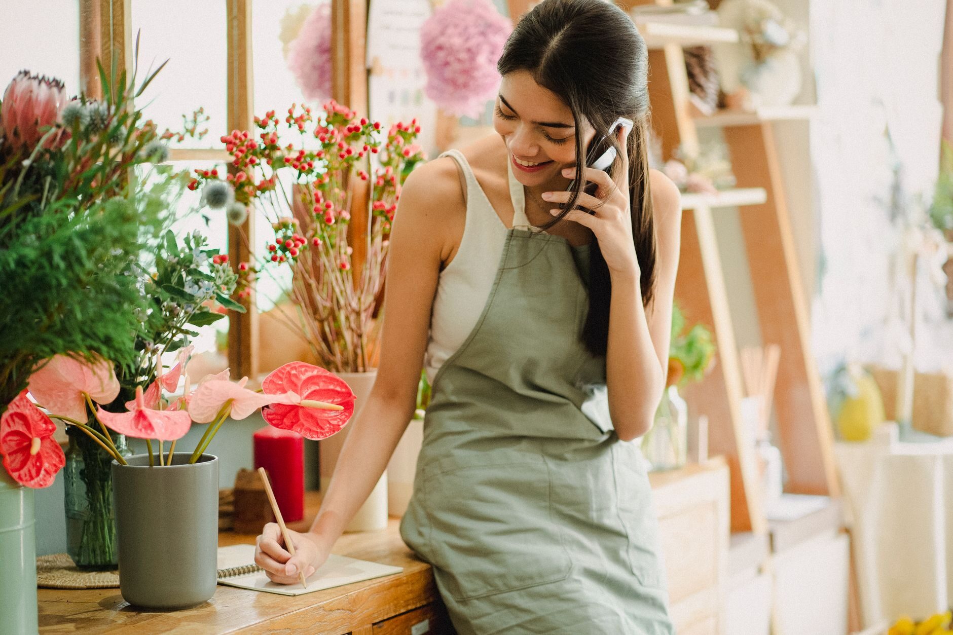 A flower-shop owner smiling while talking on her phone and writing a note at her counter, surrounded by fresh pink and red blooms