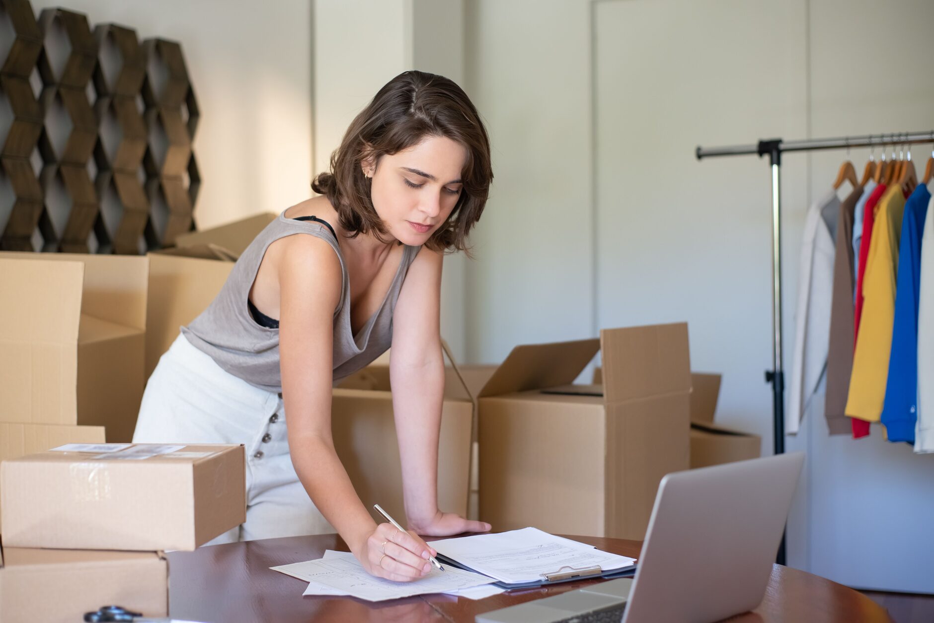 A boutique owner filling online orders at her laptop, surrounded by shipping boxes and a clothing rack